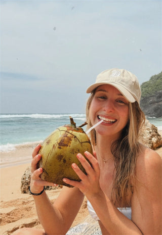 Woman on a beach holding a coconut with a straw, wearing a cap.